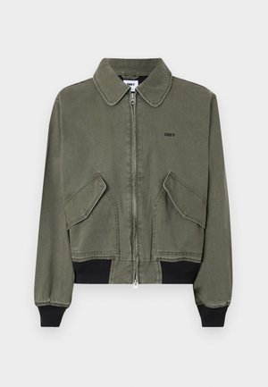 Chaqueta corta de color verde oliva con cierre de cremallera en la parte delantera, dos bolsillos frontales y puños de canalé negros. Presenta un cuello con punta y el logotipo de la marca en el pecho.