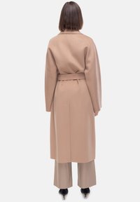 Cappotto lungo in misto lana color marrone chiaro con cintura a nastro, maniche larghe e taglio dritto, abbinato a pantaloni beige a gamba larga.