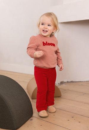 Bambino che indossa una felpa rosa con la scritta rossa "bloom", abbinata a pantaloni di velluto a coste rossi e scarpe beige, in piedi su un pavimento in legno.