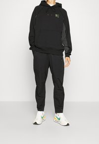 Person som bär en svart Nike Air hoodie, svarta Nike byxor och vita Nike sneakers med gröna och gula accenter, stående på ett ljust golv.