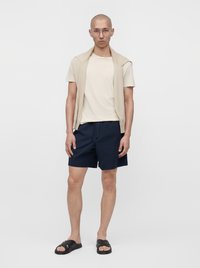 Homme portant un t-shirt beige avec un pull beige posé sur les épaules, un short bleu marine, des sandales noires et des lunettes transparentes, debout devant un fond blanc.