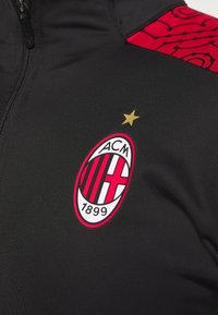 Black athletic jacket featuring an embroidered red and white AC Milan logo with a gold star above it. Textured fabric with a contrasting red sleeve detail.