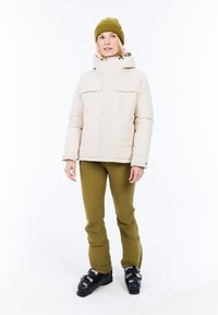 Vrouw in een beige winterjas, olijfgroene broek, mosterdgekleurde gebreide muts en zwarte skiboots, staande op een witte achtergrond.