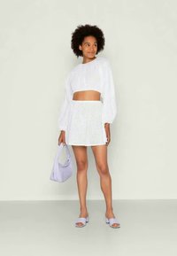 Top crop branco com mangas bufantes e saia mini branca de renda. A modelo segura uma bolsa lavanda e usa sandálias slide lavanda a condizer.