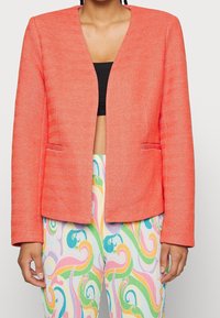 Blazer corail avec un motif chevrons texturé, devant ouvert et manches longues. Comprend deux poches avant, associé à un pantalon coloré à motifs.