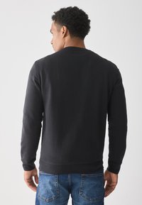 Man with short curly hair facing away wearing a black long-sleeve shirt and blue jeans against a plain white background.