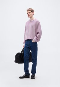 Ανοιχτό μωβ πλεκτό πουλόβερ με ριμπ λαιμόκοψη, σε συνδυασμό με μπλε τζιν. Το μοντέλο κρατά μια μαύρη τσάντα duffel, φορά μαύρα παπούτσια.