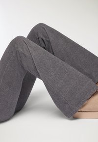 Szare rozszerzane spodnie z miękkiego, teksturowanego materiału, z widocznymi szwami i kieszeniami, zestawione z beżowym obuwiem na jasnym tle.