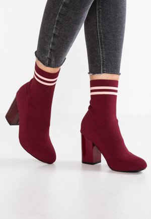 Bordowe skarpetkowe botki na słupku z różowymi paskami, noszone z czarnymi, ściętymi dżinsami na białej podłodze.