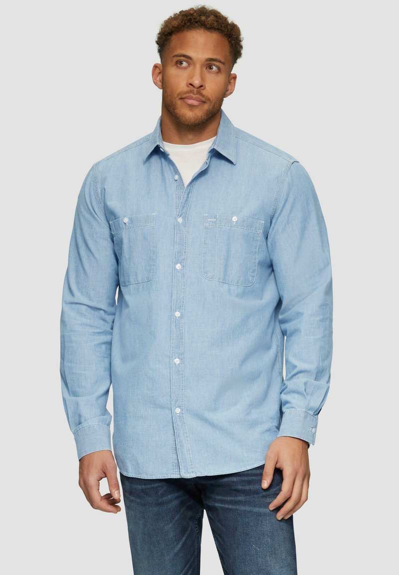 Lichtblauw denim shirt met lange mouwen, knoopsluiting, twee borstzakken en witte knopen; de stof heeft een gladde, lichtjes gestructureerde afwerking.