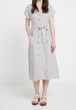 Vrouw draagt een witte midi-jurk met zwarte verticale strepen, knopen aan de voorkant, korte mouwen, een geknoopte taille en zakken, gecombineerd met witte sandalen.