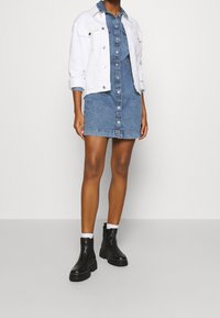 Džínové šaty s knoflíkovým zapínáním vpředu, s užším pasem a roztrhaným lemem, kombinované s bílou zkrácenou bundou a černými kotníkovými botami.