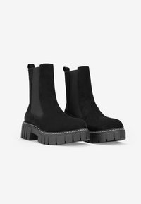 Botas de tobillo de ante negro con suela de goma gruesa, paneles laterales elásticos y tiradores para facilitar su uso. Presentan una superficie texturizada y acentos cosidos.