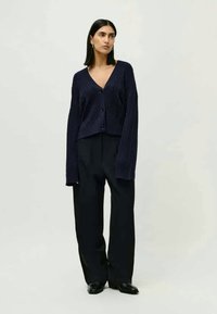 Marineblauer Strickcardigan mit V-Ausschnitt, langen Ärmeln und großen Knöpfen, kombiniert mit schwarzen, weit geschnittenen Hosen und Loafers.