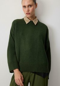 Pulover tricotat verde închis, cu o cămașă bej cu guler dedesubt. Prezintă mansete și tiv ribelate, o croială lejeră și un design asimetric.