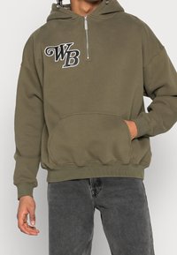 Sudadera con cremallera de color verde olivo, con un bolsillo frontal, logo bordado "WB" en el pecho, tela suave y puños y dobladillo acanalados.
