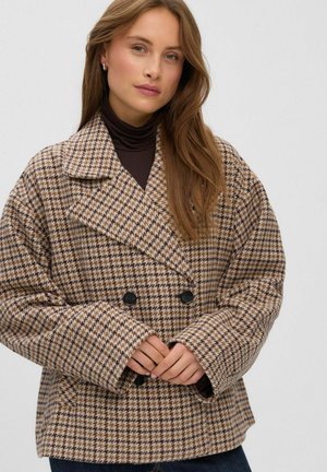 Le mannequin pose devant un fond uni, portant un manteau pied-de-poule beige et marron sur un col roulé foncé, les mains jointes.