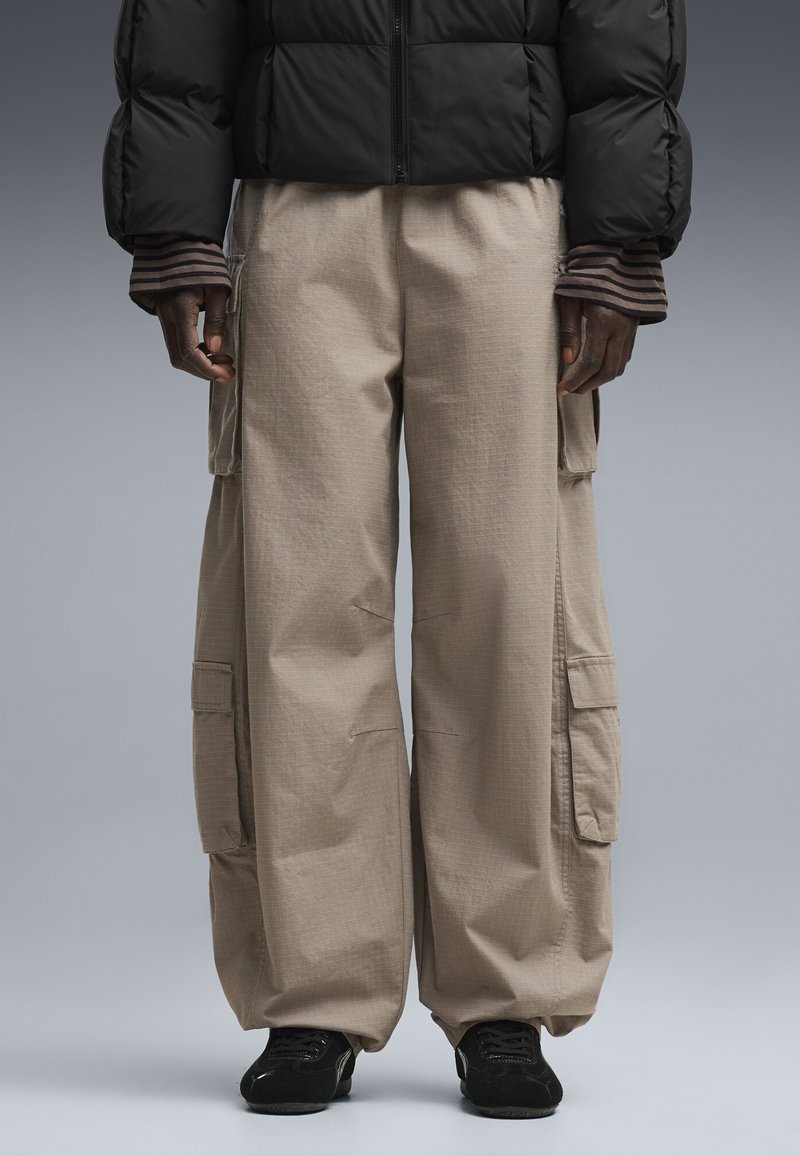Person wearing beige cargo pants, black sneakers, black puffer jacket, and striped sleeves standing against neutral background.