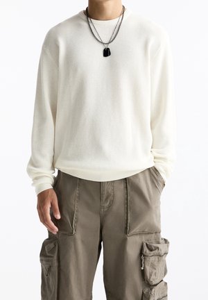 Homme portant une chemise blanche texturée à manches longues, un pantalon cargo beige avec de grandes poches et des colliers en chaînes noirs superposés. Mains détendues le long du corps.