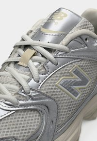Ανδρικό παπούτσι για τρέξιμο με γκρι επάνω μέρος από δίχτυ, ασημένιες συνθετικές λεπτομέρειες, κρεμ κορδόνια και εμφανές λογότυπο 'N' σε ασημί και κίτρινο χρώμα.