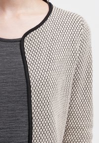 Cárdigan beige texturizado con un patrón de panal, con ribetes negros a lo largo de los bordes, superpuesto a una parte superior gris oscuro.