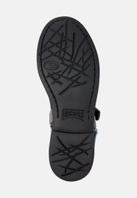 Suela de zapato de goma negra con patrones de textura, con la marca "CAMPER" y bordes curvados. Parte superior de tela gris visible en el talón.