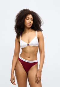 Balts trīsstūrveida krūšturis ar plānām siksnām un logotipa detaļām. Bordo tanga ar baltu elastīgu jostu, uz kuras ir teksts "TOMMY HILFIGER". Gluda auduma virsma.