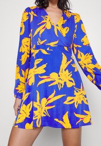 Robe bleue à effet portefeuille, manches longues, avec un motif floral jaune et rose. Tissu léger à texture lisse.