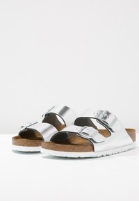 Sølv to-bånd sandaler med metalspænder, korkfodbed og hvid gummisål; glat overflade og afrundet form.