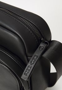 Black synthetic crossbody bag featuring a prominent zipper, textured accents, and an adjustable strap with embossed branding detail.
