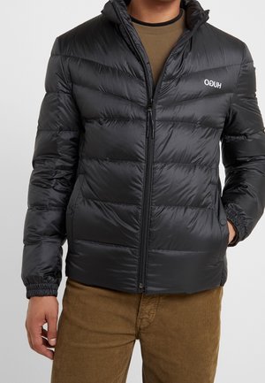 Black puffer jacket with horizontal stitching, high collar, and zip closure. Features elastic cuffs and logo on the chest. Worn over brown pants.