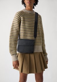 Svart krokodilpräglad axelväska med lock, i kombination med en randig tröja och en veckad kaki kjol, uppställd mot en vit bakgrund.