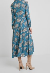 Kvinde iført en lang teal kjole med et blomstret paisleymønster og beige ruskindsstøvler med høje hæle, stående foran en ensfarvet lys baggrund.