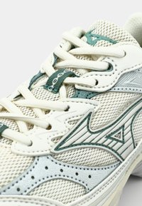 Zapatilla deportiva blanca y verde con parte superior de malla y cuero, cordones redondos y un diseño lateral distintivo. Suela texturizada y cuello acolchado.