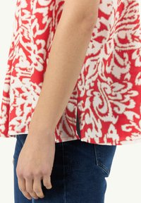 Rotes und weißes gemustertes Oberteil mit lockerer Passform, kurzen Ärmeln und seitlichen Schlitzen, kombiniert mit blauen Jeans, das eine Nahaufnahme des Materials und Designs zeigt.