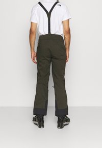 Gröna skidbyxor med justerbara svarta remmar, utrustade med förstärkta vrister och rak passform. Bärs med svarta skidkängor.