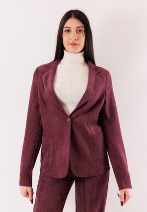Blazer in velluto a coste color bordeaux con rever a lancia, chiusura con un solo bottone e due tasche frontali. Indossato sopra un dolcevita bianco a coste.