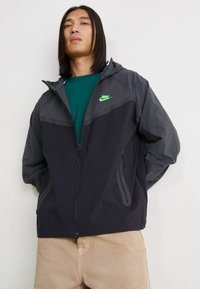 Nike jakna v odtenkih sive z zelenim logotipom. Ima kapuco, zadrgo in stranske žepe. Izdelana je iz lahkega materiala, športna zasnova.