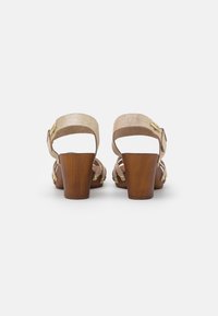Beige suede heeled sandals with a wooden block heel, open back, and adjustable ankle strap featuring gold hardware accents.