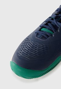 Chaussure de sport bleue avec dessus en maille texturée, semelle en caoutchouc verte et lacets. Présente un design épuré et un motif de traction distinct sur la semelle.