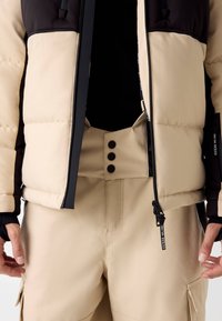 Beige pufferjack met zwarte accenten, voorzien van een hoge kraag, ritssluiting en drie zwarte knopen op bijpassende beige broek.