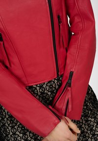 Veste en cuir rouge courte avec des zips noirs et des détails de poches. Texture lisse contre une jupe en dentelle noire à motifs.