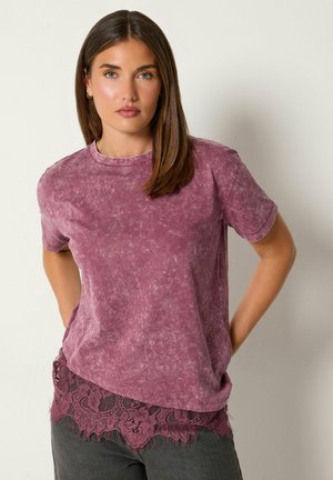 Femme aux cheveux bruns et lisses portant un t-shirt violet délavé par-dessus un haut à dentelle et un pantalon sombre, debout les mains dans le dos.