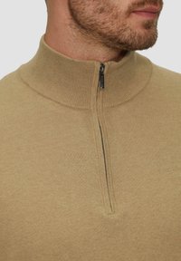 Pull en beige avec col montant en côtes et texture lisse. Il présente une fermeture éclair courte et une coupe ajustée, idéale pour le superposition.