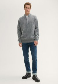 Grauer Strickpullover mit Halbzipp-Kragen, kombiniert mit blauen Jeans und schwarzen Stiefeletten. Die Ärmelsäume sind leicht gerafft.