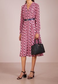 Kwiatowa sukienka midi w odcieniach czerwieni i różu, z długimi rękawami, dekoltem w kształcie V oraz dopasowaną talią z niebieskim paskiem. Całość dopełniają czarna torebka i sandały na obcasie.