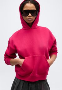 Γυναίκα που φοράει φωτεινό ροζ φούτερ με κουκούλα, με τα χέρια στην εμπρόσθια τσέπη και υπερμεγέλη μαύρα γυαλιά ηλίου, μπροστά σε απλό φόντο.