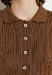 Brun ribbad stickad polo med krage och fem små ljusa knappar framtill. Strukturert tyg med vertikala linjer.