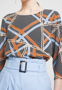 Multifarvet bluse med geometrisk mønster i sort, orange og blå, parret med højtaljede lyseblå bukser med rektangulær spænde.