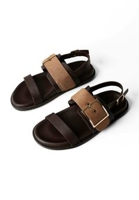 Dunkelbraune Ledersandalen mit breiten, hellbraunen Riemen, die große goldene Schnallen aufweisen, offene Zehen und verstellbare Knöchelriemen, dargestellt vor weißem Hintergrund.